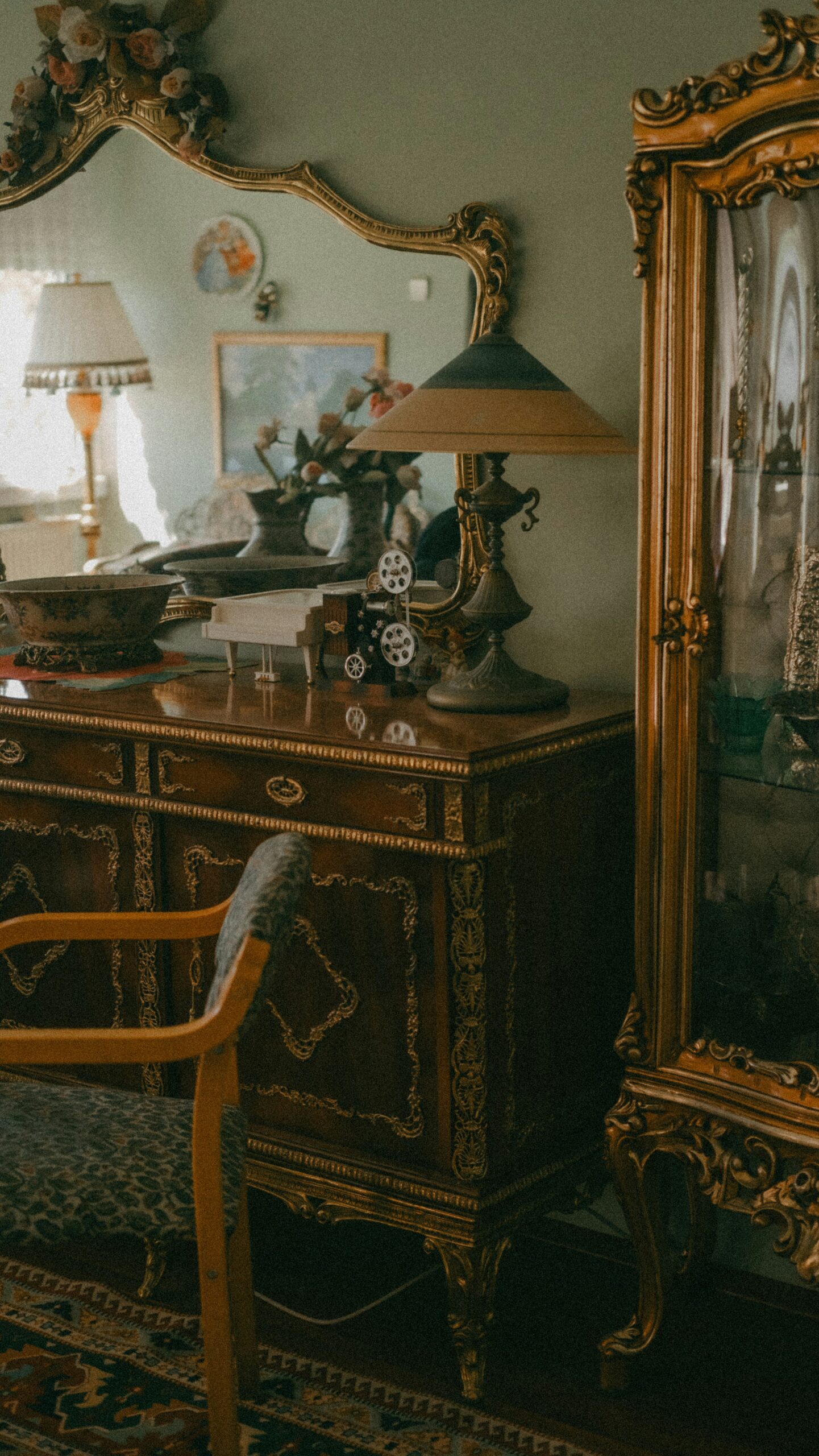 A vertical shot of a classic, antique-filled room with sage green walls. A large, gold-framed ornate mirror sits atop a dark wood sideboard decorated with gold filigree. On the sideboard are a vintage lamp with a tiered shade and a small film projector. To the right, a tall, matching gold display cabinet is visible.
